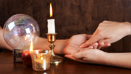 A photo of 2 people's hands together while one person gives the other a palm reading.  A candle and crystal ball sit nearby.